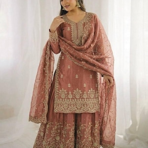 May include: A dusty rose Sharara suit featuring detailed gold embroidery. The outfit comprises a long-sleeved tunic, wide-legged trousers, and a sheer dupatta. The embroidery is focused on the neckline, cuffs, and hem of the garment.