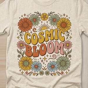 Puede incluir: Camiseta de color beige claro con un diseño floral retro. La camiseta muestra las palabras "COSMIC BLOOM" en una fuente groovy, rodeadas de flores coloridas, estrellas y un sol. El diseño es en tonos naranja, amarillo, rosa y azul.