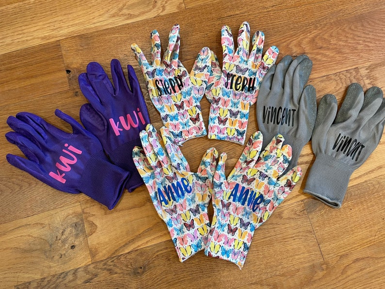 May include: A collection of gardening gloves in various designs. Two pairs are solid purple with the name "kwi" in pink. Two pairs feature a butterfly pattern with names "Steph" and "Anne". Two gray pairs have the name "Vincent".