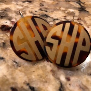 May include: A pair of gold monogram earrings with a tortoiseshell pattern. The earrings are round and have the letters "SHP" in the center.