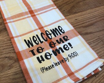 May include: A white and orange plaid kitchen towel with the text "Welcome to our home! (Please leave by 9:00)" printed in black.