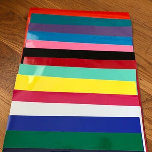 May include: A stack of colorful vinyl sheets in various shades, including red, teal, purple, blue, pink, black, red, mint green, yellow, magenta, white, and navy. The sheets are arranged in a neat, organized pile.