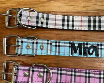 May include: Three dog collars with plaid patterns. The top collar is white with black and red plaid. The middle collar is light blue with black and white plaid and the name "Mia" printed on it. The bottom collar is pink with black and white plaid.