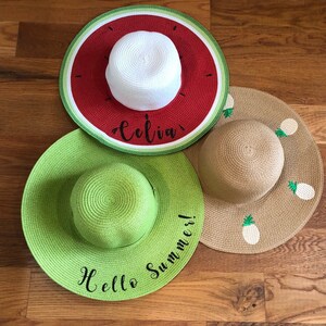 May include: Three straw sun hats with wide brims. One hat is red and green with a watermelon design and the name "Celia". Another is light green with the text "Hello Summer!". The third hat is tan with pineapple designs.