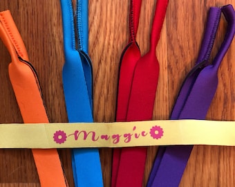 May include: Five colorful eyeglass straps with black edging. The straps are arranged in a rainbow pattern, with orange, blue, red, and purple straps. A yellow strap with the word "Maggie" printed on it is positioned across the middle of the straps.