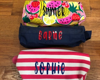 May include: Three fabric pencil cases with different designs. The top case is pink with a watermelon, pineapple, lemon, and banana pattern and the word "summer" printed on it. The middle case is navy blue with the name "barne" printed in red. The bottom case is red and white striped with the name "sophie" printed in blue.