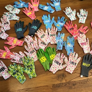 May include: A collection of gardening gloves in various colors and patterns, including floral and polka dot designs. Many gloves are personalized with names like "Jess," "Suzie," and "Linda." Some gloves are solid colors, such as green and gray.