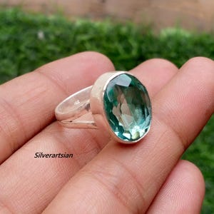 May include: A silver ring with a large, oval-shaped, light teal gemstone. The ring has a split band design. The text "Silverartsian" is visible on the image.