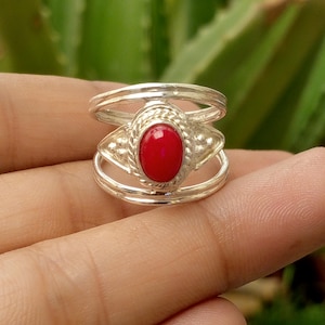 May include: A silver ring featuring a central oval red stone set within a twisted metal border. The band consists of two parallel silver wires that connect to the central setting, which is accented with small silver spheres on each side.