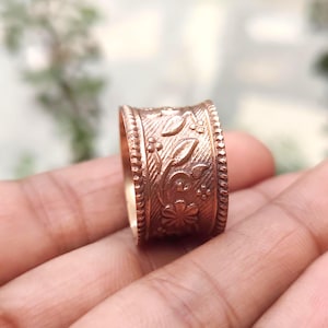 Pode incluir: Um anel largo em tom de cobre com um padrão floral e de folhas em relevo na banda central. As bordas do anel são definidas por uma textura de contas em relevo que emoldura o intrincado design botânico gravado na superfície metálica.