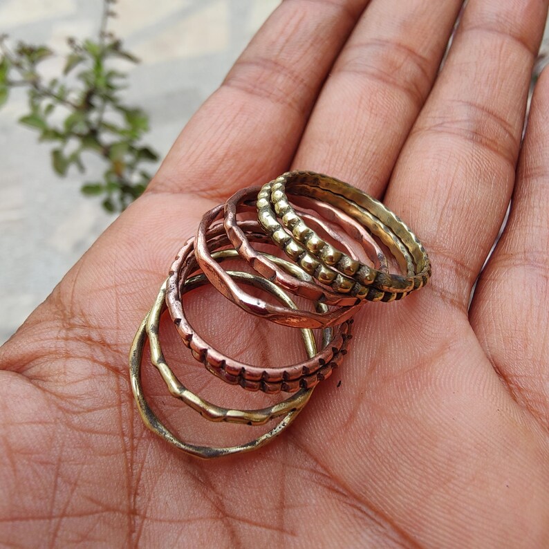 May include: A selection of six brass and copper rings in various designs. The rings are stacked, displaying different textures and patterns, including beaded and wavy details. The rings are presented on a hand.