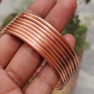 May include: A set of multiple stacked copper-colored bangles featuring a smooth, rounded, and polished metallic surface. The bracelets are thin, circular, and uniform in design, creating a reflective and shiny appearance across the entire set.