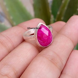 May include: A silver ring featuring a faceted, pear-shaped, vibrant pink gemstone set in a smooth metal bezel. The word Silverartsian is printed in black text on the band. The ring has a polished silver band and a pointed, teardrop-shaped stone.