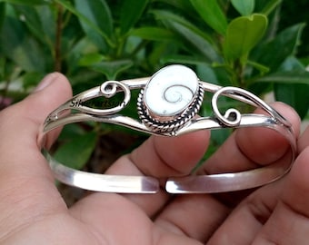 May include: A silver cuff bracelet featuring a central oval-shaped white shiva eye shell with a spiral pattern. The metal band has a polished finish with decorative swirling wire details on both sides of the stone setting, which is framed by a twisted rope border.