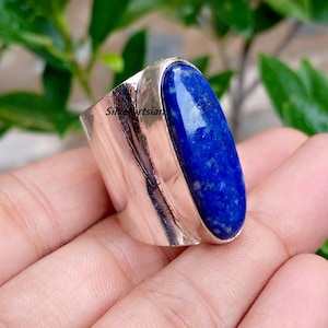 May include: A wide silver ring featuring a large, oval-shaped, deep blue lapis lazuli gemstone set vertically. The polished metal band has a smooth, reflective surface with the text Silverartsian printed clearly on the side of the metal setting.