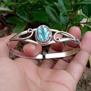 May include: A silver cuff bracelet featuring an oval-shaped abalone shell inlay at the center. The shell displays iridescent green, blue, and purple tones. The metal band has a polished finish with decorative scrollwork and a twisted rope border.
