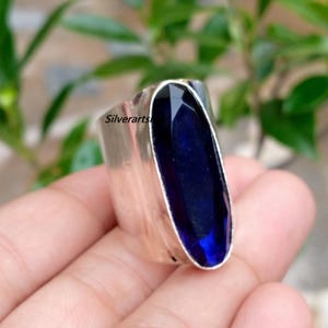 May include: A wide silver ring featuring a large, elongated, faceted deep blue gemstone set vertically. The metal band has a smooth, polished finish. The text "Silverarts" is visible on the side of the silver band near the gemstone setting.