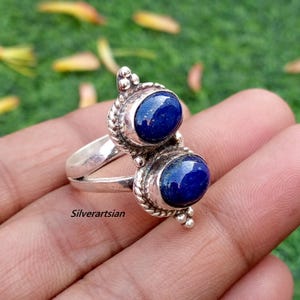 May include: A silver ring featuring two oval, deep blue lapis lazuli stones. The ring has a decorative, textured border around the stones and a split shank. The text "Silverartsian" is visible on the ring.