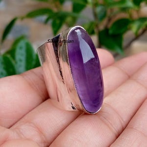 May include: A wide silver ring featuring a large, smooth, oval-shaped purple amethyst gemstone set in a polished metal band. The word Silverartsian is printed in small black text on the side of the silver band, which has a reflective surface.
