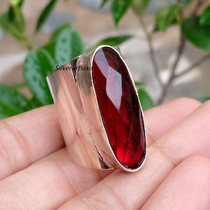 May include: A wide silver ring featuring a large, elongated, faceted deep red gemstone set vertically. The metal band has a smooth, polished finish and the text Silverartsian is visible on the side of the setting. The design is bold and metallic.