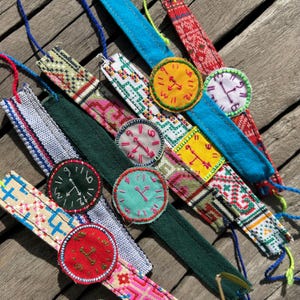 Puede incluir: Una colección de coloridas correas de reloj de tela hechas a mano con esferas bordadas. Las correas presentan varios patrones y colores, incluyendo azul, verde, rojo y amarillo. Cada esfera de reloj tiene un color diferente y muestra una hora diferente.