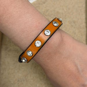 May include: A narrow orange leather bracelet featuring a row of six round, clear, faceted crystals set in silver-toned metal bezels. The band has a smooth texture and a dark edge, secured with a simple closure visible at the end of the strap.