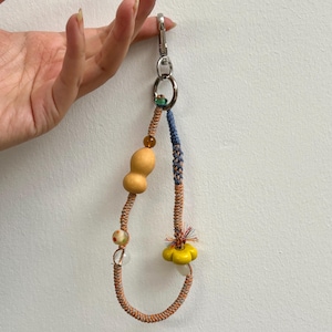 May include: A handmade keychain with a silver clasp and ring. The keychain features a gourd-shaped wooden bead, colorful beads, and a yellow flower-shaped bead. The cord is a mix of orange, blue, and brown.