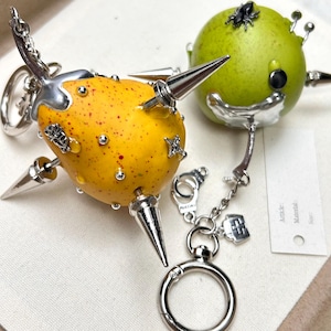 Peut inclure: Deux porte-clés fantaisie. L'un est jaune avec des pointes et des ornements argentés. L'autre est vert avec des accents argentés. Les deux ont des porte-clés et des breloques argentés. Le porte-clés jaune a des mouchetures rouges.