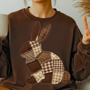 May include: A brown sweatshirt with a patchwork rabbit design in shades of brown and beige. The rabbit is made from different fabric patterns, including stripes, gingham, and floral prints. The sweatshirt is a plain brown colour.