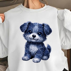 以下が含まれることがあります： 白いスウェットシャツ。青いデニムパッチワークデザインの子犬の漫画が特徴です。子犬は大きな青い目を持ち、笑顔の表情をしています。スウェットシャツはカジュアルなスタイルで、普段使いに適しています。