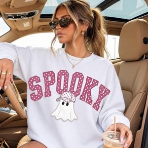 May include: A white long-sleeve sweatshirt featuring the word SPOOKY in large, pink, polka-dotted varsity-style letters. Below the text is a graphic of a white ghost wearing a matching pink polka-dotted baseball cap, centred on the chest.