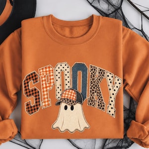 Puede incluir: Una sudadera naranja de cuello redondo con la palabra SPOOKY en letras grandes de estilo universitario con patrones variados que incluyen cuadros naranjas, lunares negros, mezclilla y estampado de leopardo. Un fantasma blanco con gorra está debajo.