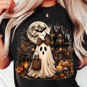 May include: A black t-shirt featuring a graphic print of a white ghost wearing a leopard print bow, holding a matching leopard print bag. The scene includes a full moon, bats, a haunted house, orange autumn leaves, lanterns, a pumpkin, and a skull.
