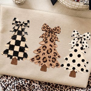 Puede incluir: Tres recortes decorativos de árboles de Navidad con troncos marrones, cada uno rematado con un lazo grande. El primer árbol presenta un patrón de cuadros negros y crema, el segundo un estampado de leopardo beige y marrón, y el tercero lunares negros.