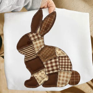 Può includere: Un articolo in tessuto bianco con un design a coniglio patchwork. Il coniglio è composto da vari tessuti marroni, tra cui pezzi a tinta unita, a righe e fantasia. Il coniglio è in posizione seduta con le orecchie dritte.