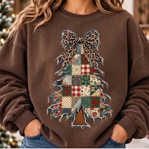 May include: A brown crewneck sweatshirt featuring a large graphic of a Christmas tree made of various patchwork fabric patterns. The tree is topped with a leopard print bow and has frayed, distressed edges throughout the design.