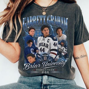 May include: A charcoal gray t-shirt featuring a graphic print of hockey player Garrett Graham. The design includes multiple images of the player, the text "GARRETT GRAHAM", "Briar University", "HOCKEY TEAM", and "#44" with blue lightning accents.