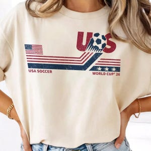 Może przedstawiać: Kremowa koszulka z grafiką USA Soccer. Projekt zawiera czerwone litery "US", piłkę nożną oraz napis "USA SOCCER WORLD CUP '26" w kolorze granatowym i białym. Na koszulce znajduje się r&oacute;wnież flaga amerykańska.