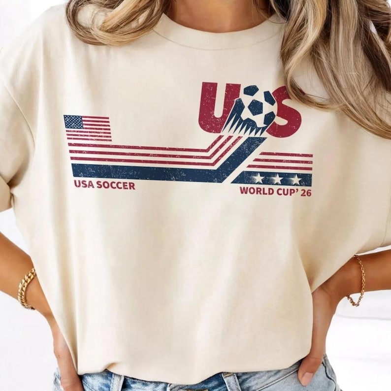 Może przedstawiać: Kremowa koszulka z grafiką USA Soccer. Projekt zawiera czerwone litery "US", piłkę nożną oraz napis "USA SOCCER WORLD CUP '26" w kolorze granatowym i białym. Na koszulce znajduje się r&oacute;wnież flaga amerykańska.