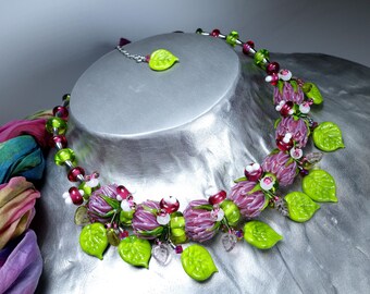 May include: A handcrafted necklace featuring a floral design with purple, green, and red glass beads. The necklace includes leaf-shaped pendants and a matching earring. The jewelry is displayed on a silver bust.