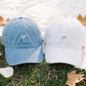 Puede incluir: Dos gorras de béisbol, una en azul vaquero desteñido y otra en blanco sólido, cada una con un pequeño icono de palmera bordado en blanco o azul claro en el panel frontal, con visera curva y un diseño estructurado de seis paneles.