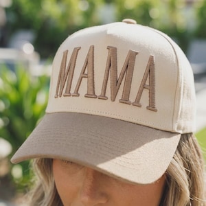 May include: A beige baseball cap featuring the word MAMA embroidered in brown thread on the front panel. The cap has a structured crown and a solid brown brim, with a button at the top centre and visible stitching along the brim and panels.