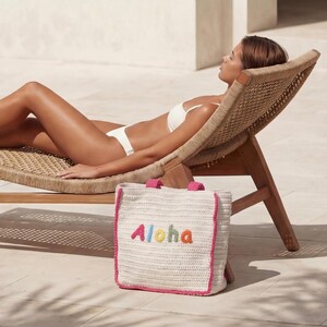 Op de afbeelding: Een rechthoekige, gebroken witte gehaakte tas met het woord Aloha in veelkleurige letters op de voorzijde geborduurd. De tas heeft een felroze rand langs de zijkanten en twee bijpassende roze hengsels en staat rechtop op een lichte ondergrond.