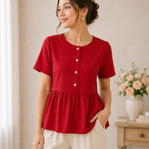 Può includere: Un top rosso a maniche corte con scollo tondo e bottoni bianchi. Il top ha un dettaglio a peplo in vita. La modella indossa pantaloni beige. Il tessuto è leggero.