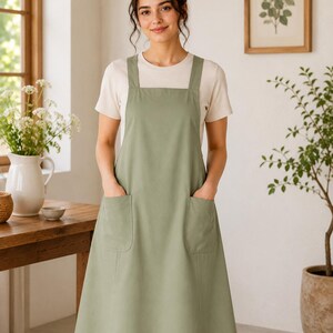 May include: A sage green apron with wide shoulder straps and two large front patch pockets. The apron features a straight neckline and a knee-length skirt, worn over a plain short-sleeved cream-colored t-shirt made of a solid fabric.
