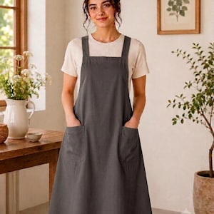 May include: A charcoal gray pinafore apron featuring a square neckline, thick shoulder straps, and two large front patch pockets. The apron is worn over a short-sleeved, light beige crew-neck t-shirt, creating a layered look with a simple silhouette.