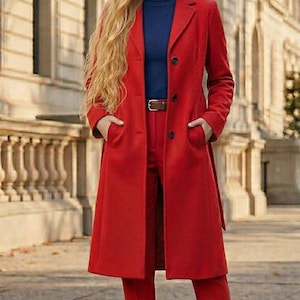 Puede incluir: Un abrigo de lana rojo vibrante hasta la rodilla, usado sobre un suéter de cuello alto azul oscuro y pantalones rojos a juego. El abrigo presenta solapas, tres botones negros y dos bolsillos laterales. Se ve un cinturón de cuero marrón.