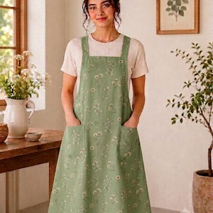May include: A sage green pinafore apron featuring a delicate white floral pattern, a square neckline, thick shoulder straps, and two large front patch pockets. The apron is worn over a plain, short-sleeved, light beige crew-neck t-shirt.