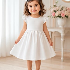 May include: A young child wearing a white dress with ruffled sleeves and a flared skirt. The dress is paired with white shoes. A white bow is in the child's hair. The background includes a table with flowers and a mirror.