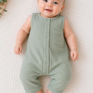 May include: A smiling baby wearing a sage green sleeveless romper with a front pocket and button closure. The baby is lying on a white, textured blanket. The romper is made of a soft, textured fabric.